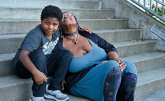 A young Black boy and his mom sit on concrete steps. His arm is thrown around her shoulder and he smiles into the camera. Her eyes are closed, and she leans back onto his shoulder.