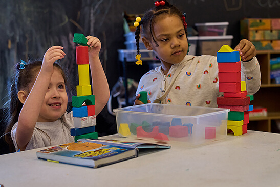 Two young girls play with colorful blocks on a white table. There's a book open in front of them, and behind them is a chalkboard.