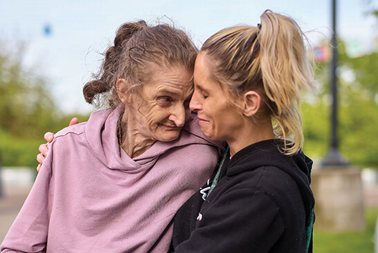 An elderly woman with light-brown hair in a ponytail and wearing a pink hoodie, smiles and touches foreheads with a middle-aged woman with blonde hair in a ponytail and wearing a black hoodie.