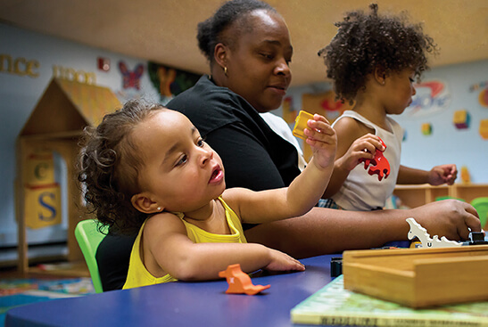 A mom with two toddlers play with dinosaur toys and books at a blue table.