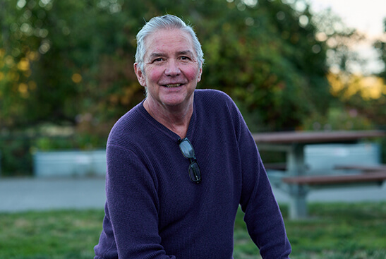 A grey-haired man wearing a navy-blue pullover smiles broadly for the camera. He sits outdoors on a bench.