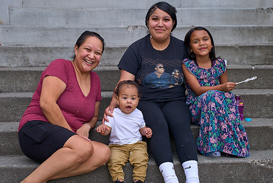 A multigenerational family sit on concrete steps, smiling. They include a grandmother, mother, young daughter, and baby boy.