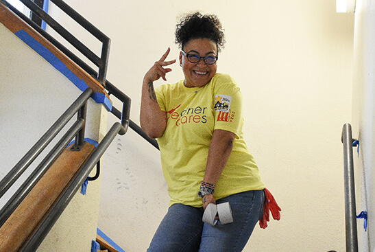 A woman in a yellow T-shirt smiles and makes a peace sign with her fingers while standing in a freshly painted stairwell.