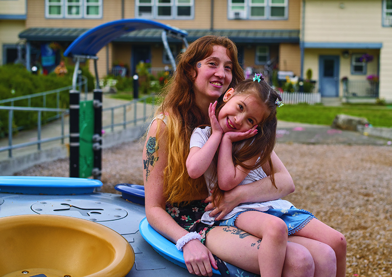 A young girl with long brown hair sits on her mother's lap in a playground with an apartment building in the background. Both are in shorts and the girls is cupping her hands around her tilted head.