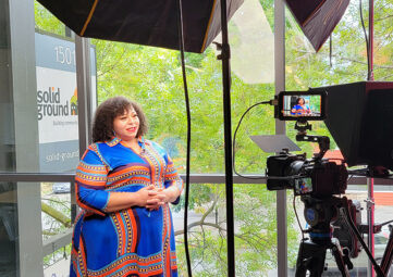 A woman with curly hair and a colorful blue and gold dress stands in front of a TV camera with a sign that reads "Solid Ground" visible through the window behind her.
