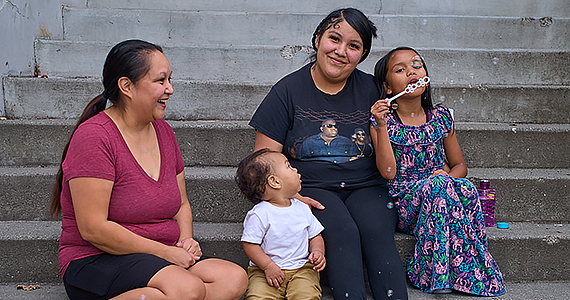 A grandmother, mom, baby boy and young daughter sit together on concrete steps and smiling. The little girls is blowing bubbles.