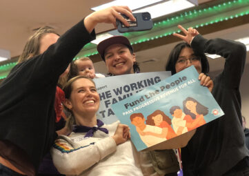 A close up shot of a group of people smiling and holding signs as one of them takes a group selfie with a phone.