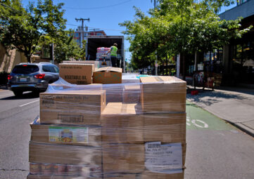 A shrink-wrapped pallet of boxed food, including vegetable oil and split peas, sits on the side of a street as a man in yellow vest off loads another pallet from the back of a truck.