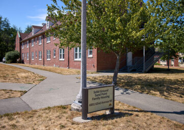 A sign that reads "Solid Ground Sand Point Family Housing" In front a long brick building with two stories.