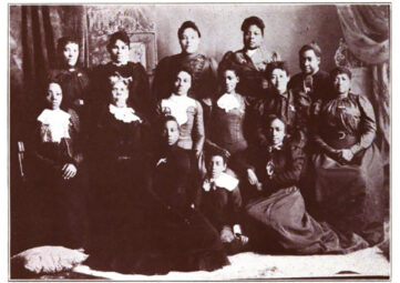 A historic black-and-white photo of about a dozen women in formal dressed posing together.
