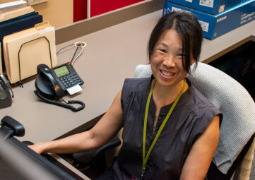 A wiling woman sits behind a computer monitor at an office desk.