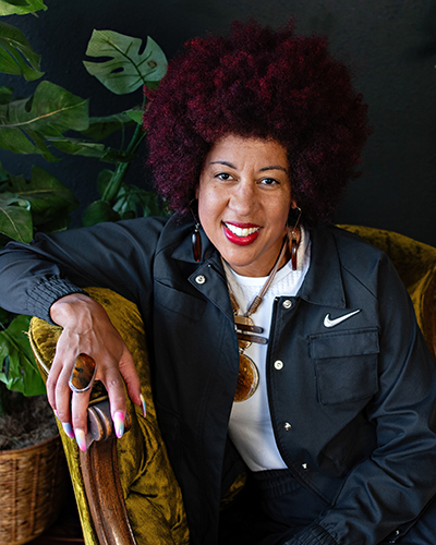 Portrait of a Black woman with a reddish afro wearing a denim jacket and white T-shirt poses in a gold armchair with green plants behind her.