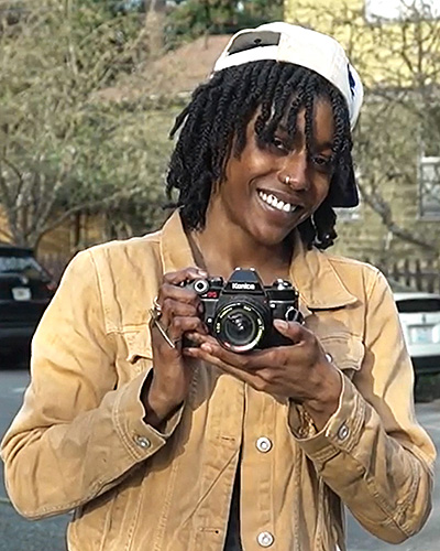 Portrait of a Black woman with short locs wearing a beige collared shirt, and holding a camera in her hands.