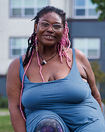 Portrait of a Black woman with long pink and black locs wearing glasses and a slate blue summer top. Behind her is multi-level housing.