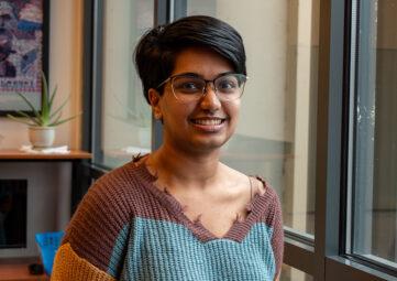 A young woman wearing a brown, orange, and blue knit-sweater poses by a wall of windows .