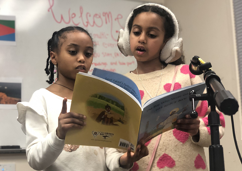 Two young girls behind a microphone reading from a picture book.