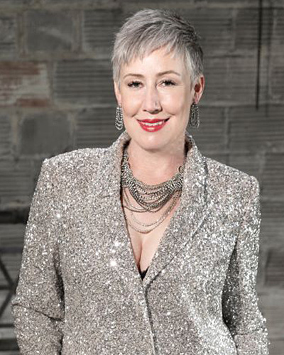 Portrait of a white woman with a short blond pixie cut, wearing a sparkly silver blazer.