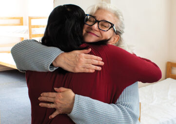 Two women hug with single beds showing in the background. The one facing away has dark brown hair and wears a red blouse. The one facing forward has grey hair and glasses, with her eyes closed, and wears a grey sweater.