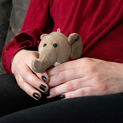 Hands of a woman holding a small stuffed elephant. She wears black nail polish, black pants, and a red blouse.