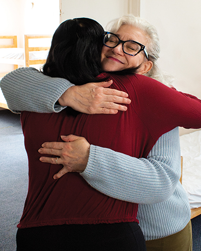 Two women hug with single beds showing in the background. The one facing away has dark brown hair and wears a red blouse. The one facing forward has grey hair and glasses, with her eyes closed, and wears a grey sweater. In the upper right corner is a blue GIVE♥BIG 2025 logo.