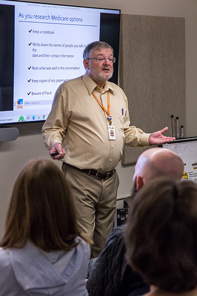 A man giving a presentation stands in front of a projection that reads "As you research medicare options," followed by five bulleted items: "Keep a notebook," "write down the names of the people you talk to, he date, and their contact information." Note what was said in the conversatons," "Keep copies of any paperwork."and "Beware of fraud."