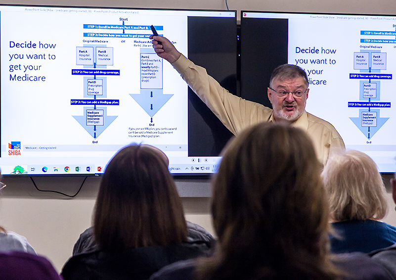 A man giving a presentation in front of a seated audience gestures at the top of a flow chart labeled "Decide how you want to get your Medicare."