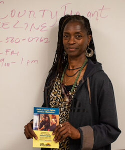 Woman with long black locs poses in front of a whiteboard with red writing on it. She holds a brochure about Tenant Services.