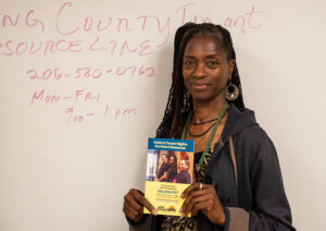 Woman with long black locs poses in front of a whiteboard with red writing on it. She holds a brochure about Tenant Services.