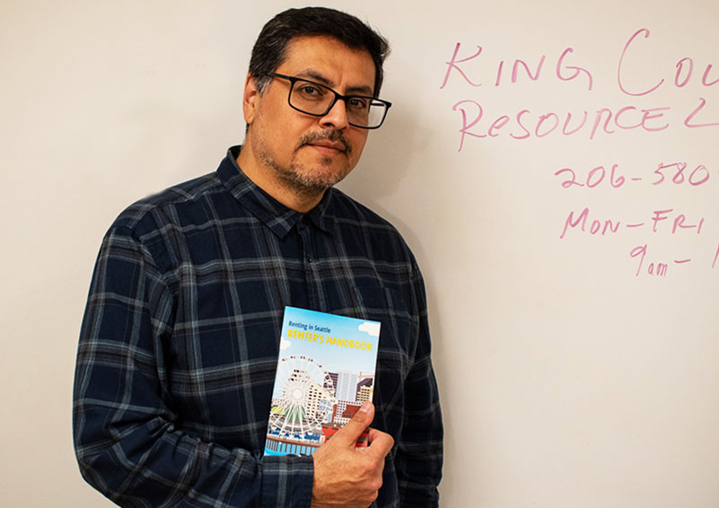 Man with dark hair and glasses, wearing a dark blue plaid shirt, poses in front of a whiteboard with red writing on it. He holds a brochure about Tenant Services.