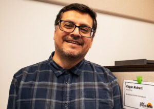 Man with dark hair and glasses, wearing a dark blue plaid shirt, poses in front of a whiteboard.