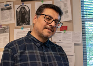 Man with dark hair and glasses, wearing a dark blue plaid shirt, poses in a work cubicle.