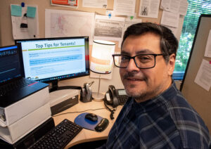 Man with dark hair and glasses, wearing a dark blue plaid shirt, poses in a work cubicle in front of a computer screen with a PowerPoint presentation up that reads, "Top Tips for Tenants!"