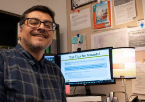 Man with dark hair and glasses, wearing a dark blue plaid shirt, poses in a work cubicle in front of a computer screen with a PowerPoint presentation up that reads, "Top Tips for Tenants!"