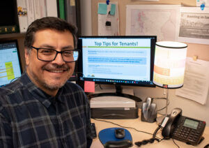 Man with dark hair and glasses, wearing a dark blue plaid shirt, poses in a work cubicle in front of a computer screen with a PowerPoint presentation up that reads, "Top Tips for Tenants!"