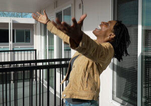 A young woman in a tan jacket stands on her apartment deck with her arms outstretched. Her head is thrown back, and she has a beaming smile on her face.
