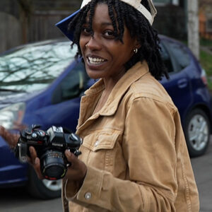 A young woman in a tan jacket and baseball cap beams as she takes photos on the move.