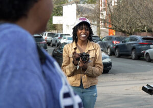 A young woman in a tan jacket and baseball cap takes photos of her mother, who stands in the photo foreground.