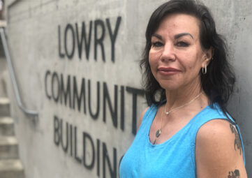 A woman with long dark hair, tattoos, and a teal tank top stands in front of a concrete wall with the words "Lowry Community Building."