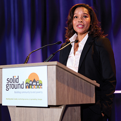 A woman in a dark suit jacket stands at a podium with a sign that reads "Solid Ground."