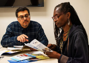 A man with dark hair and glasses and a woman with long locs and glasses sit together, talking, at a table covered with brochures and pamphlets.