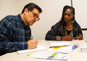 A man with dark hair and glasses and a woman with long locs and glasses sit together, talking, at a table covered with brochures and pamphlets.