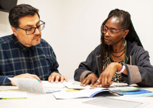 A man with dark hair and glasses and a woman with long locs and glasses sit together, talking, at a table covered with brochures and pamphlets.