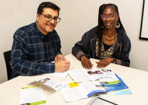 A man with dark hair and glasses and a woman with long locs and glasses sit together, smiling, at a table covered with brochures and pamphlets.