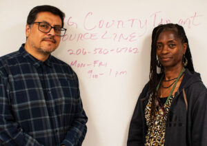 A man with dark hair and glasses and a woman with long locs pose in front of a whiteboard with red writing on it.