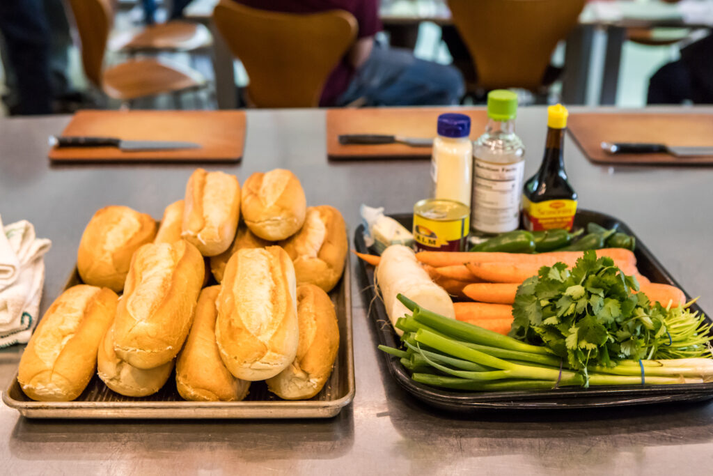 A stack of French rolls alongside carrots, scallions, and other ingredients for bahn mi sandwiches.