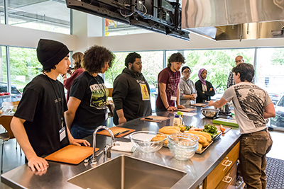 Half a dozen teenagers stand along a stainless steel kitchen countertop listing to a pair of instructors.