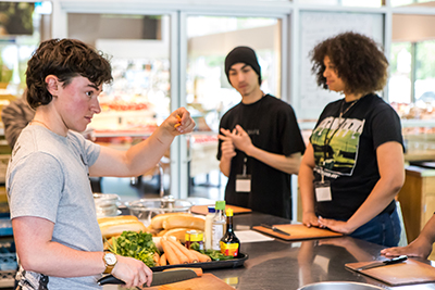 A cooking instructor standing in front of a counter piled with ingredients holds out a julienned carrot as two students look on.