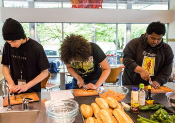 Three teenagers lean over a stainless steel countertop, carefully chopping carrots on individual wood cutting boards.