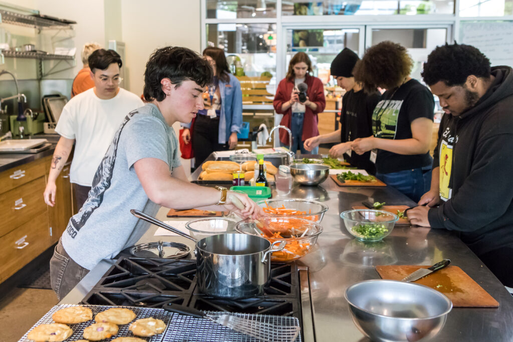 A cooking class instructor leans across a crowded kitchen counter top as students work to prepare various ingredients.