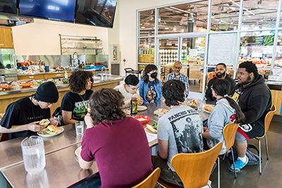 A group of students and instructors crowd around a table eating bahn mi sandwiches. A brightly lit grocery store is seen through floor-to-ceiling windows behind them.